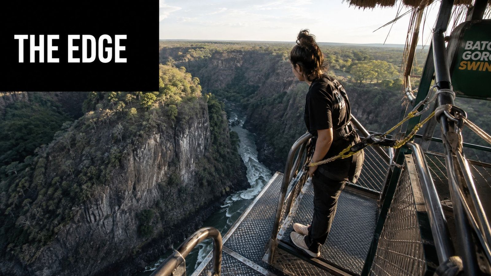 A woman stands on a metal platform looking out over the deep Victoria Falls gorge canyon.